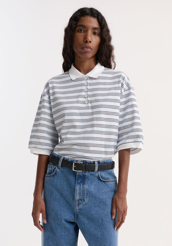 Striped oversized polo in white/blue worn by a model, featuring a four-button placket and traditional collar, paired with high-waisted jeans and a black belt.
