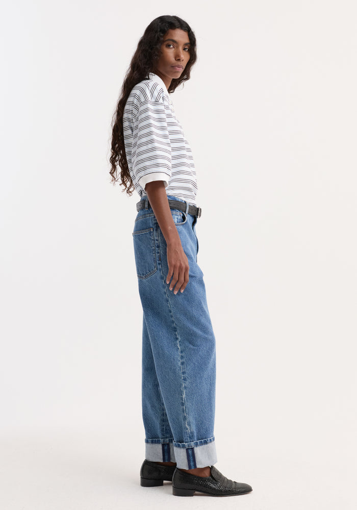 Woman wearing a striped oversized polo with a traditional collar and four-button placket, tucked into high-waisted jeans, styled with a black belt and loafers.