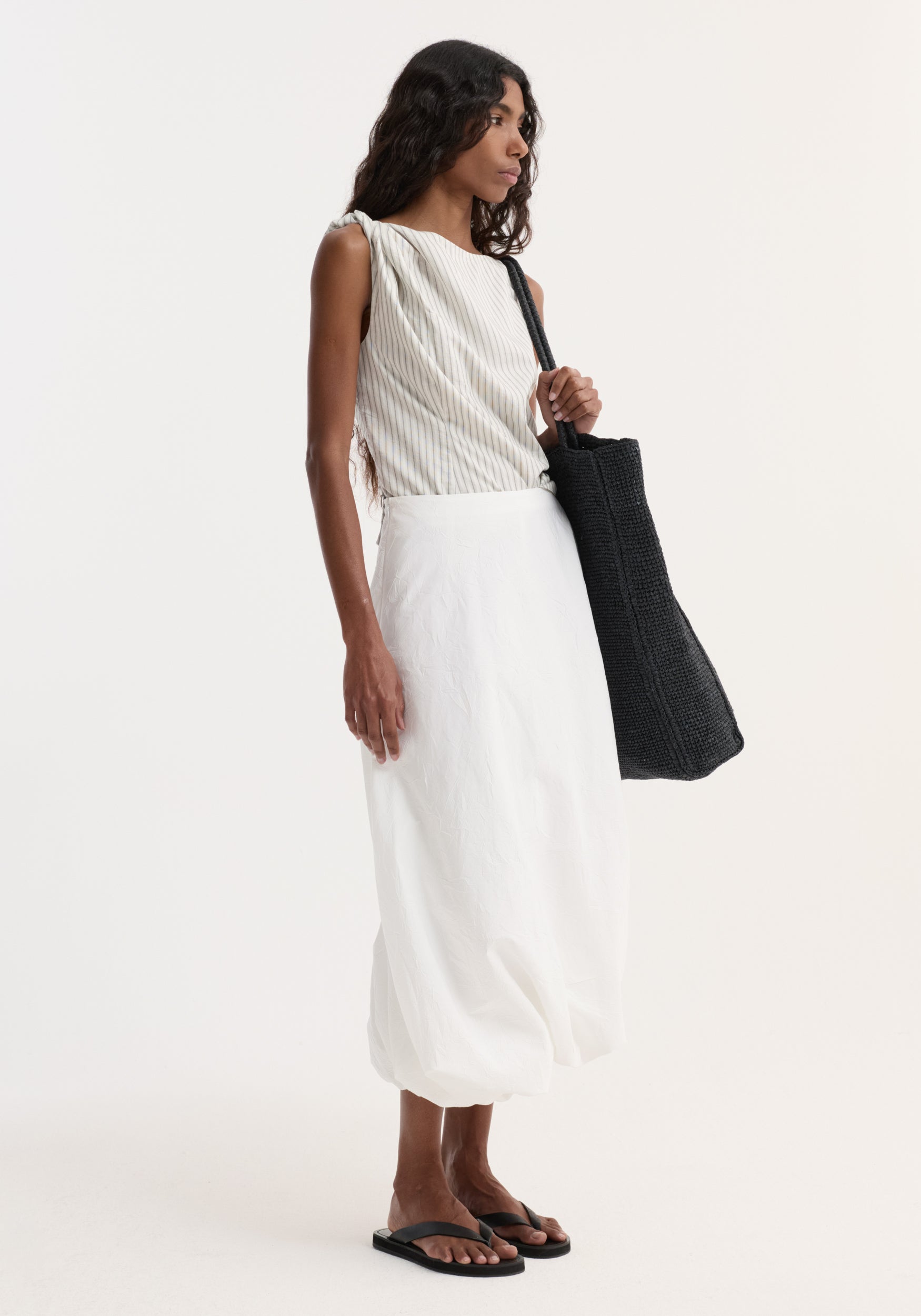 Model wears Silk-blend draped top | fine stripe with twist detail, boat neck, and gathered waist, paired with a white skirt and black tote.