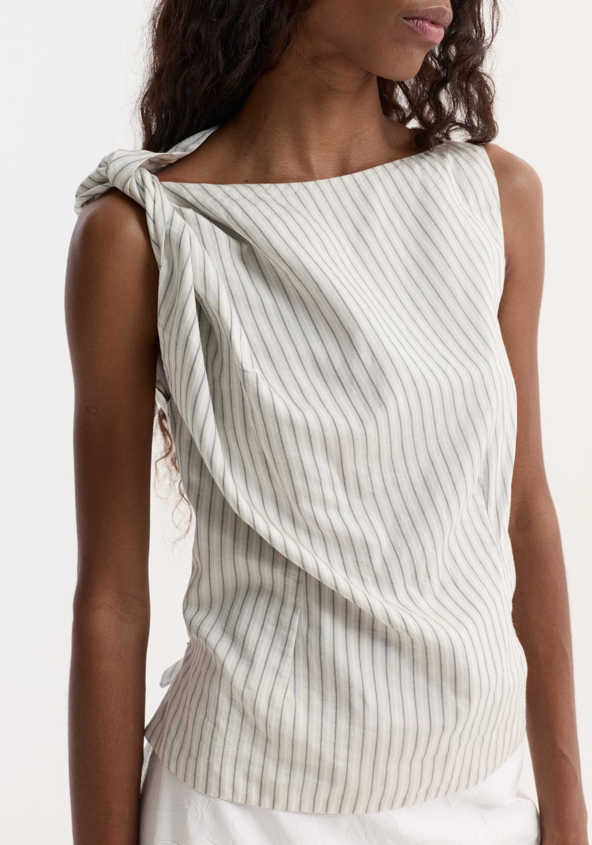 Silk-blend draped top with fine stripes, featuring a twist detail on the shoulder, boat neck, and gathered waist, worn by a slim, dark-skinned woman.