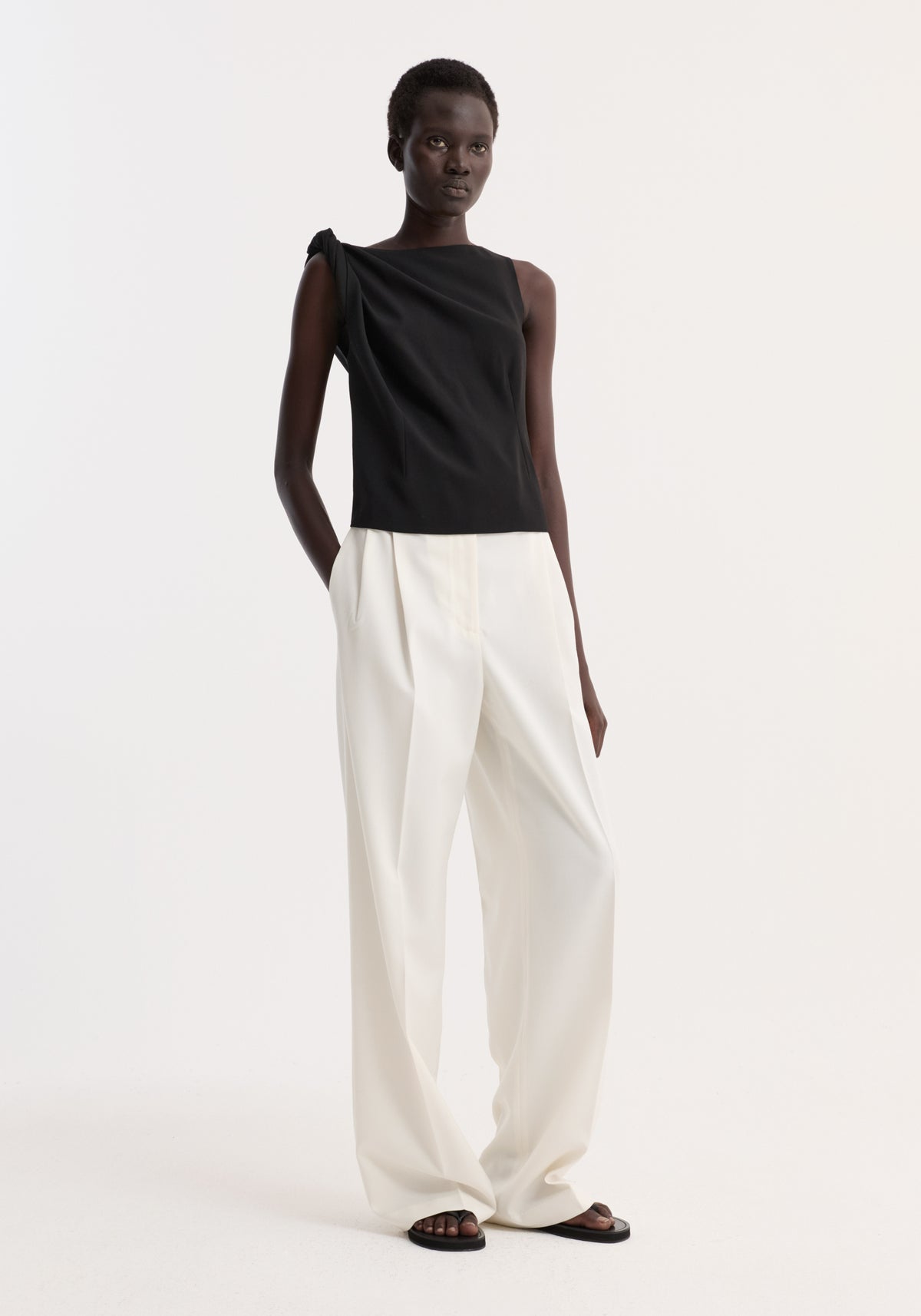 Model wears a Draped linen silk top | black, featuring a twist detail on one shoulder, boat neck, and gathered waist, paired with wide-leg trousers and flat sandals.