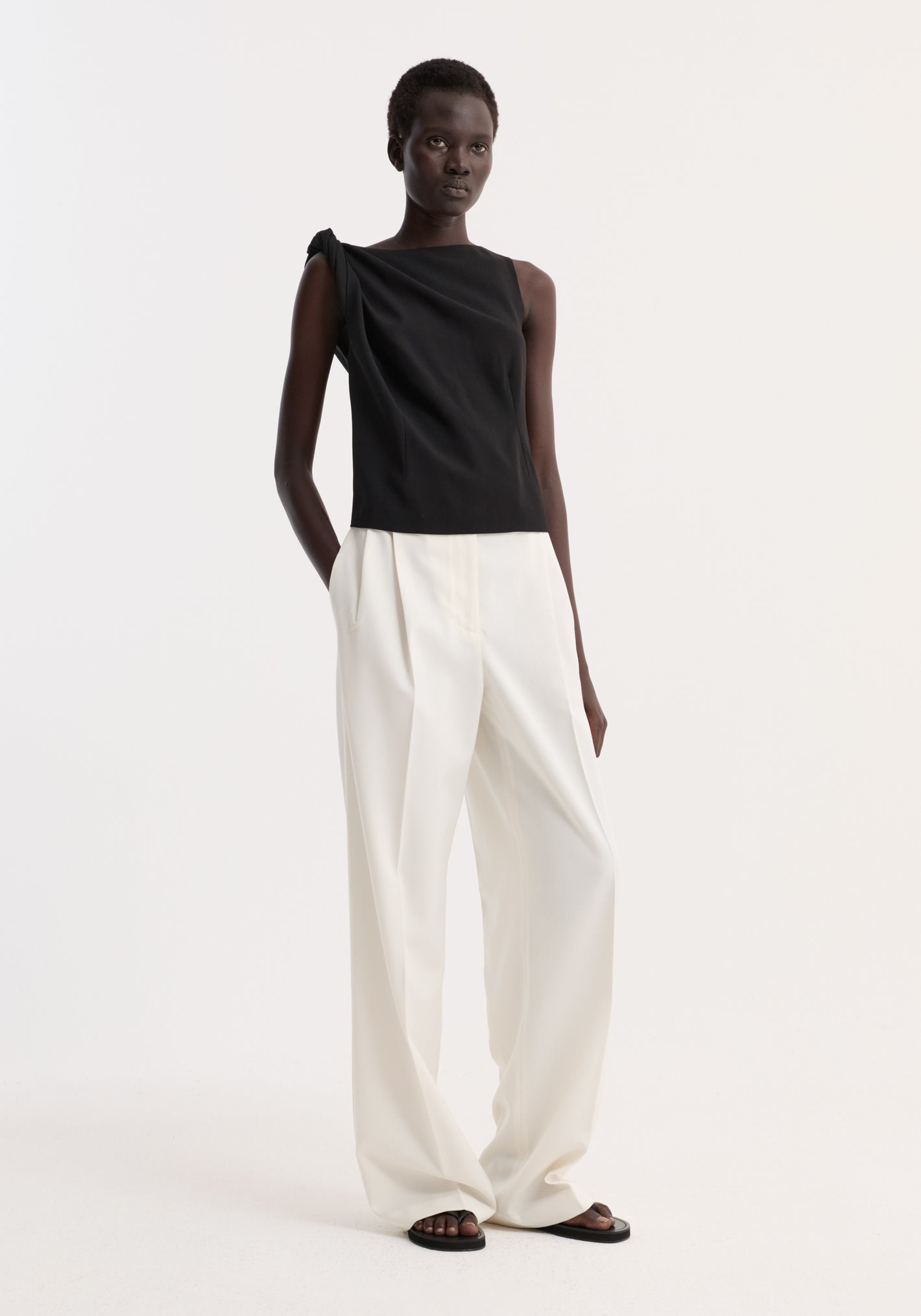 Model wears a Draped linen silk top | black, featuring a twist detail on one shoulder, boat neck, and gathered waist, paired with wide-leg trousers and flat sandals.