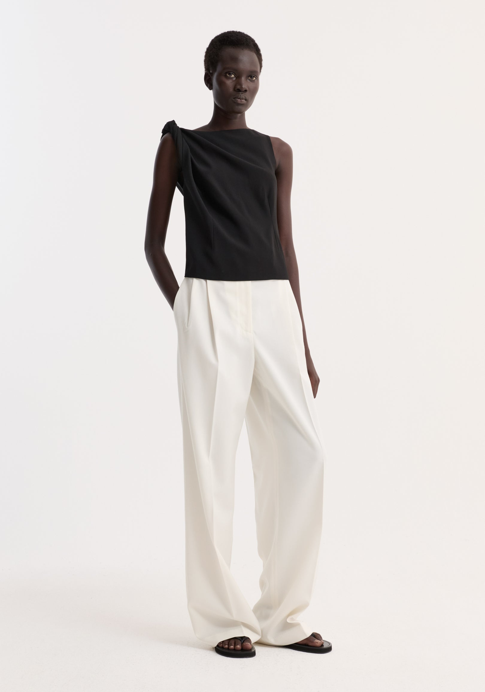 Model wears a Draped linen silk top | black, featuring a twist detail on one shoulder, boat neck, and gathered waist, paired with wide-leg trousers and flat sandals.