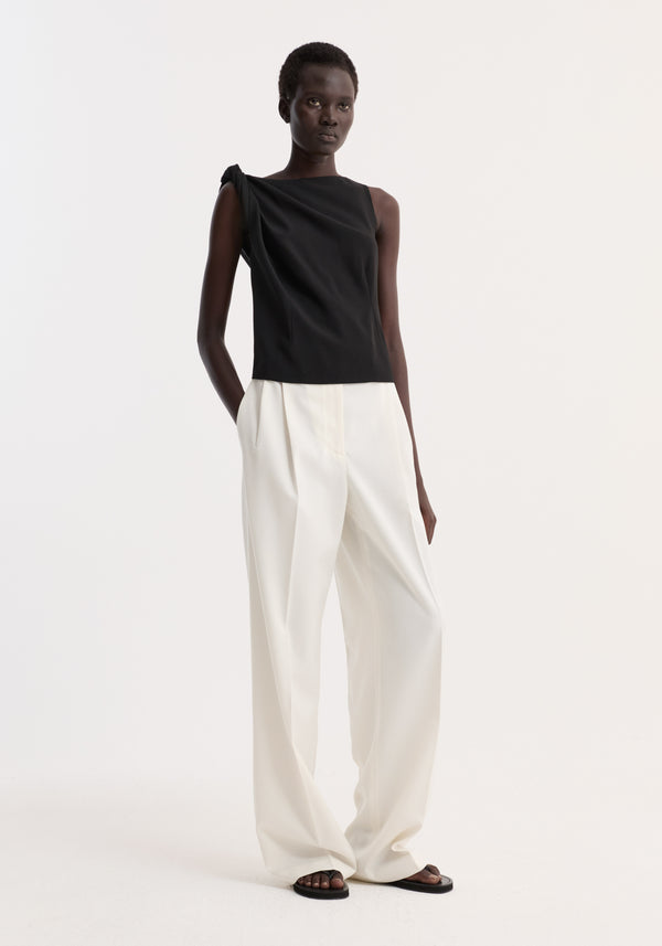 Model wears a Draped linen silk top | black, featuring a twist detail on one shoulder, boat neck, and gathered waist, paired with wide-leg trousers and flat sandals.