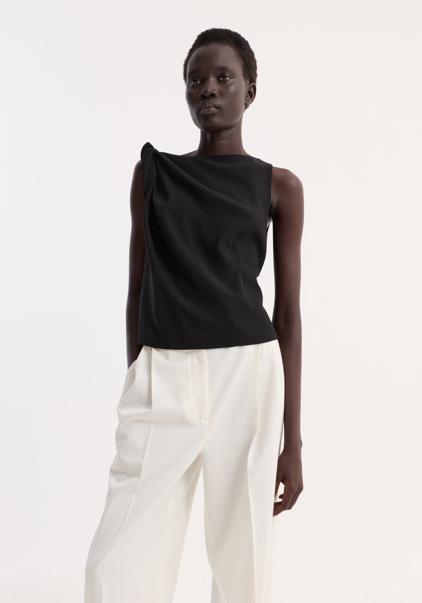 Draped linen silk top | black: Model wears sleeveless top with twist detail and asymmetrical drape, paired with high-waisted wide-leg trousers, emphasizing a sculptural silhouette.