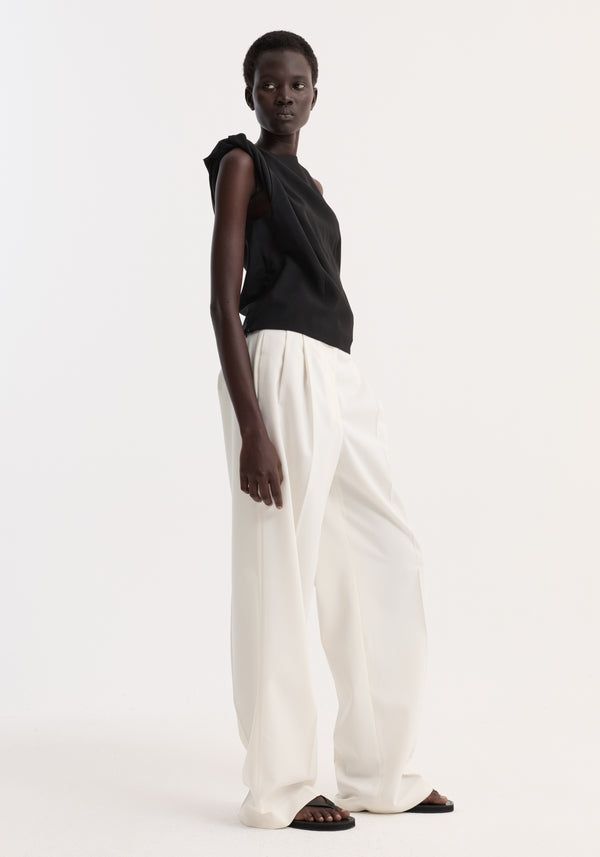 Model wearing a Draped linen silk top | black with twist shoulder detail and boat neck, paired with wide-leg trousers, showcasing a minimalist, asymmetrical design.