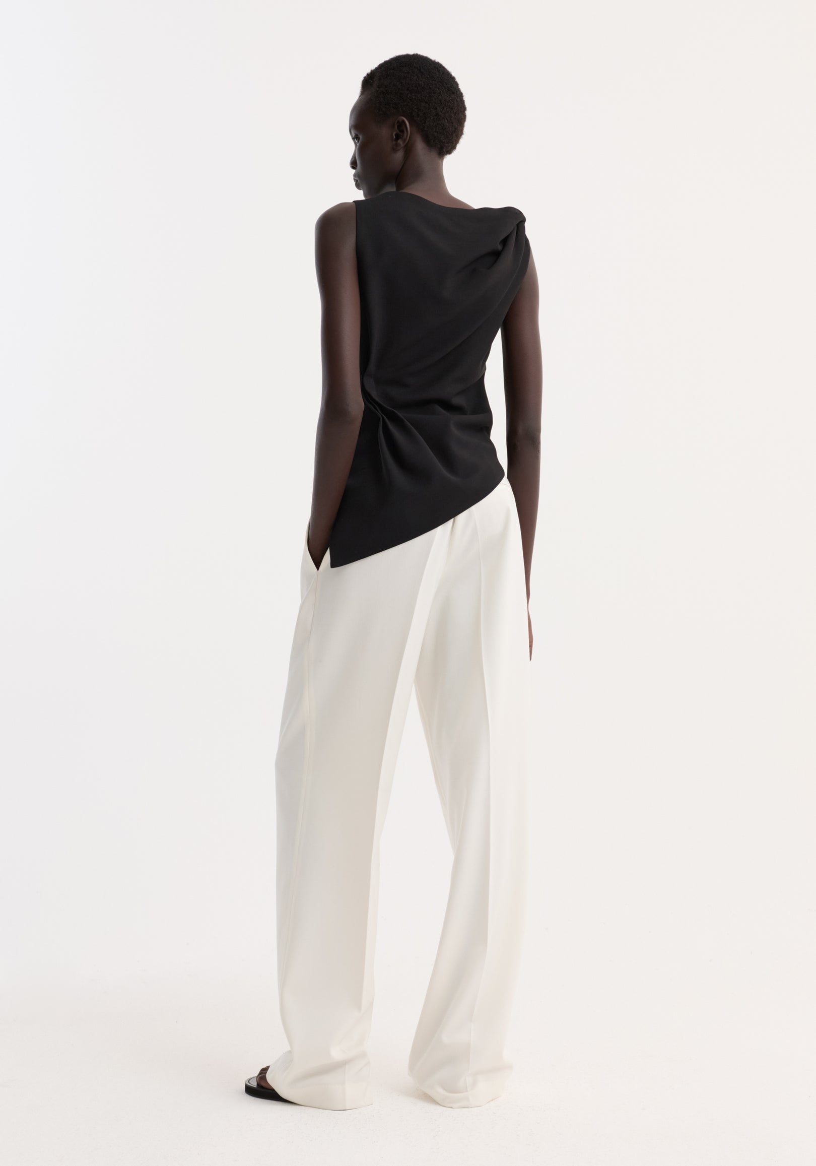 Model in Draped linen silk top | black, featuring a twist detail and boat neck, paired with high-waisted trousers, hands in pockets, showcasing an asymmetrical silhouette.