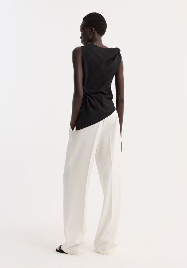 Model in Draped linen silk top | black, featuring a twist detail and boat neck, paired with high-waisted trousers, hands in pockets, showcasing an asymmetrical silhouette.