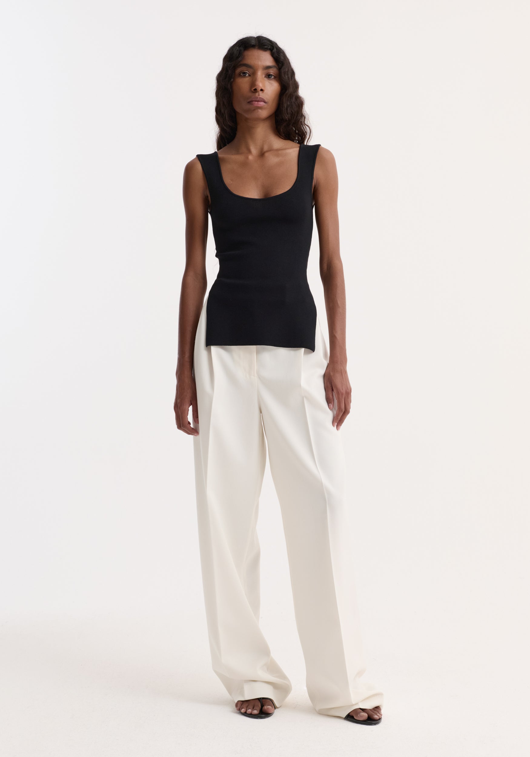 Model wearing Long knitted bustier top in black, scoop-neck, fitted design, with side splits, paired with high-waisted white trousers and black sandals.