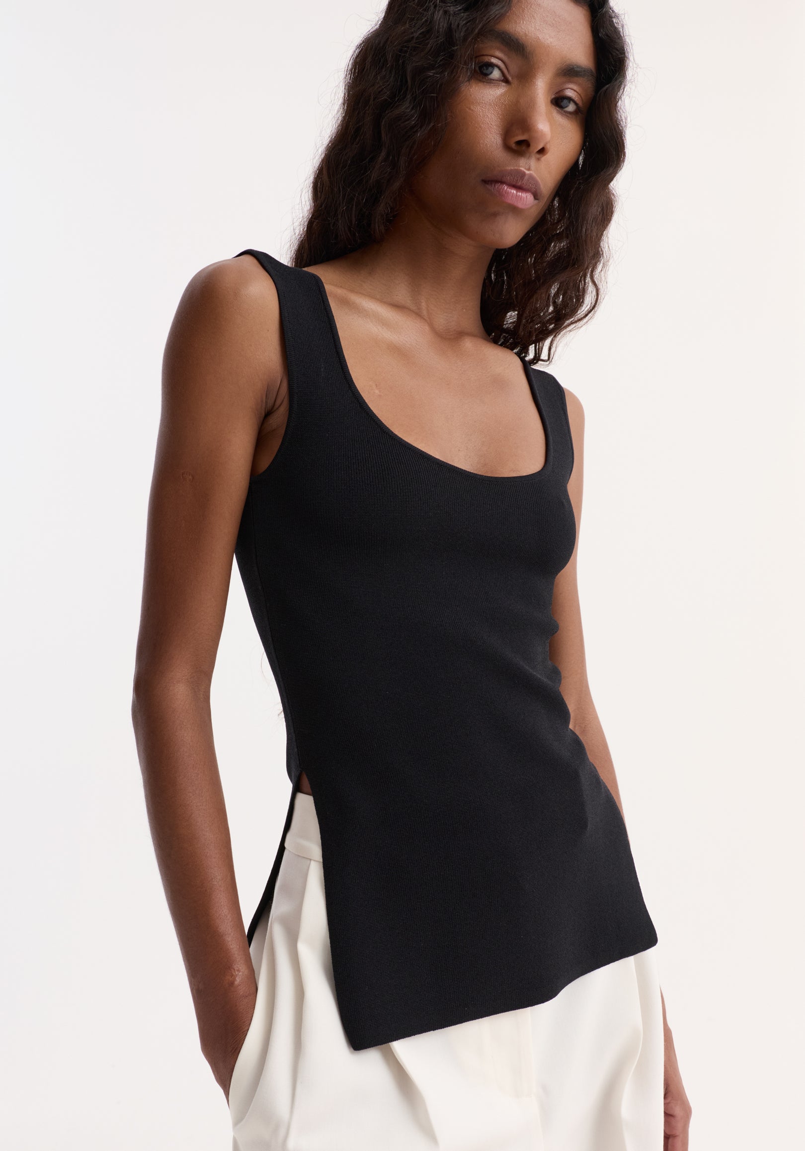 Long knitted bustier top in rib-knit with scoop neckline and side slit, worn by model with loose bottoms, showcasing fitted style and elongated silhouette.