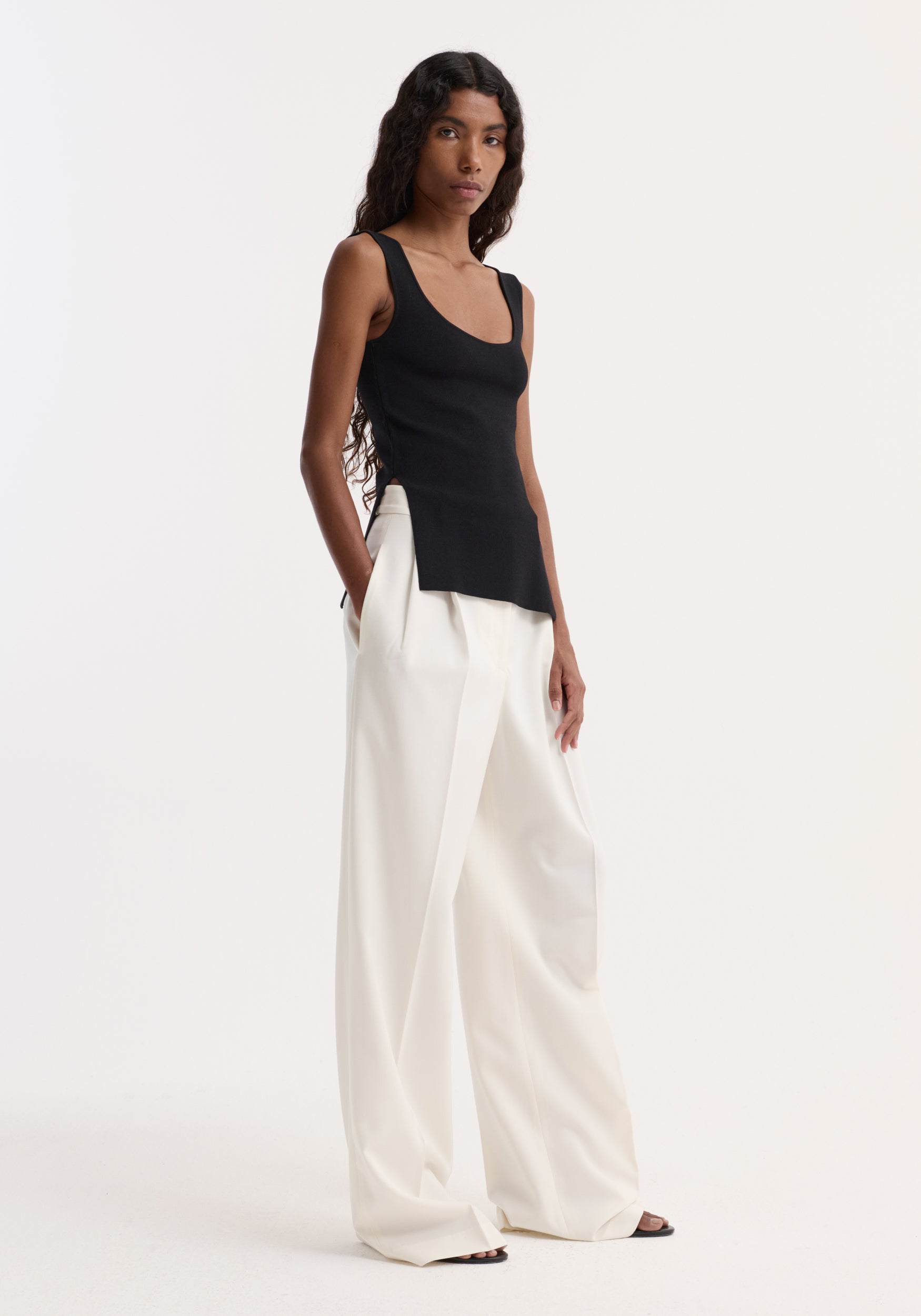 Long knitted bustier top in black, scoop neck, fitted design with side splits, worn by model with high-waisted trousers, showcasing elongated silhouette.