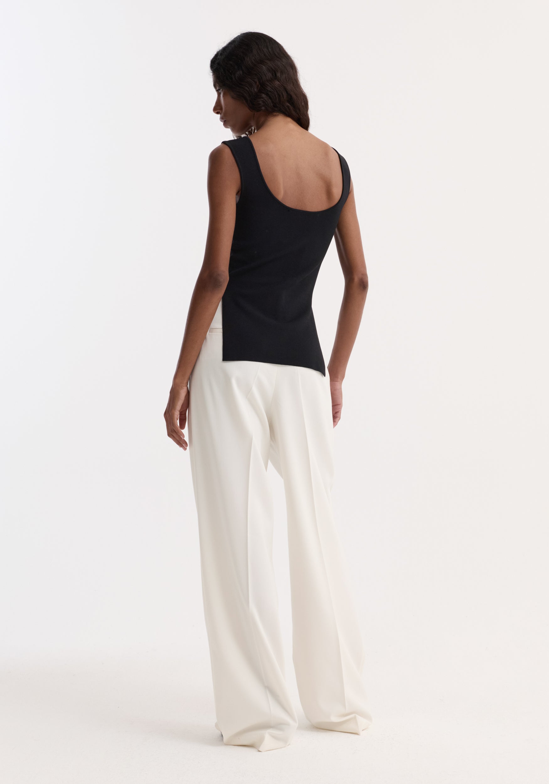 Long knitted bustier top in black, scoop neckline, fitted design, visible side splits, worn by a slim model with high-waisted, wide-leg trousers.