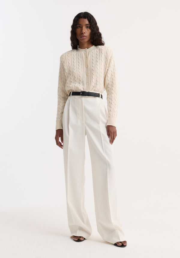 Fine merino cable cardigan in cream, with shoulder pads and a slightly cropped fit, worn with high-waisted trousers and black sandals by the model.