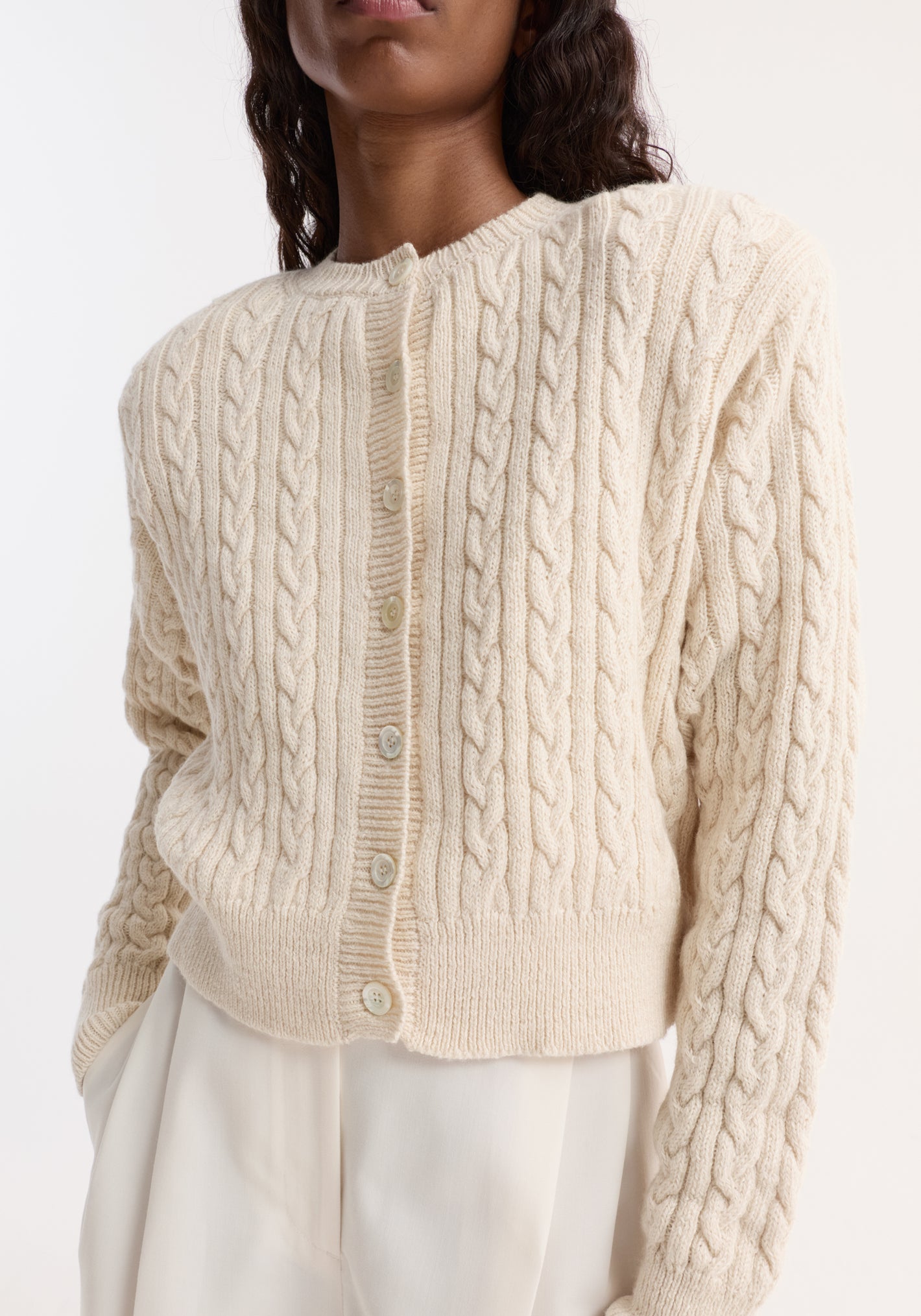 Fine merino cable cardigan in cream, featuring a textured knit with shoulder pads, small buttons, and a slightly cropped fit, worn with matching trousers.
