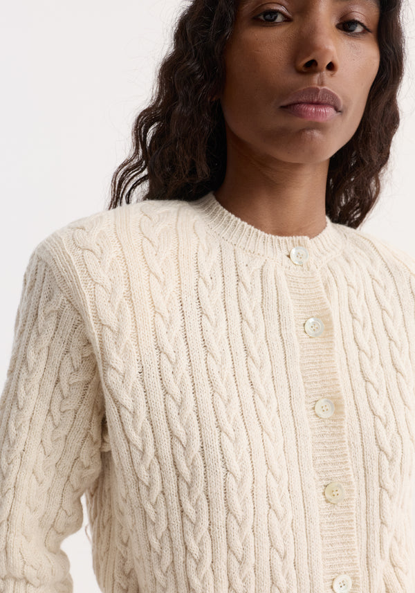 Fine merino cable cardigan in cream, showcasing textured cable-knit pattern, mother-of-pearl buttons, and subtle shoulder pads, worn slightly cropped on model’s shoulder.