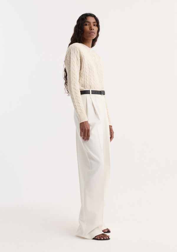 Fine merino cable cardigan in cream, slightly cropped with shoulder pads, styled with high-waisted ivory trousers and black belt. Model is 177cm, size 36.