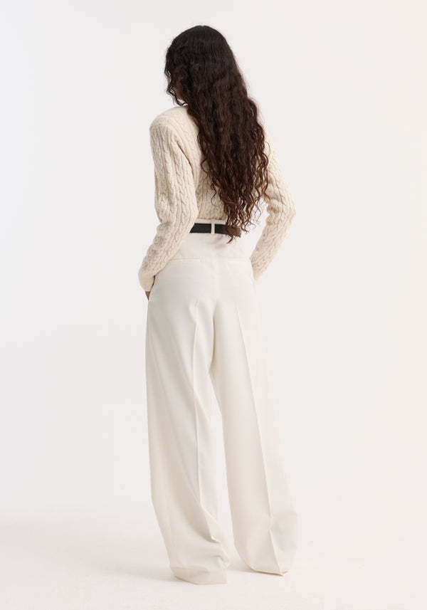 Fine merino cable cardigan in cream, slightly cropped with shoulder pads, worn tucked into high-waisted trousers. Model's hands in pockets, hair cascading down.
