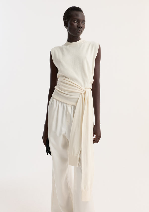 Model in open back knitted wrap top with padded shoulders, sash-style sleeve extensions, and wrap waist, paired with loose trousers, holding a black clutch.