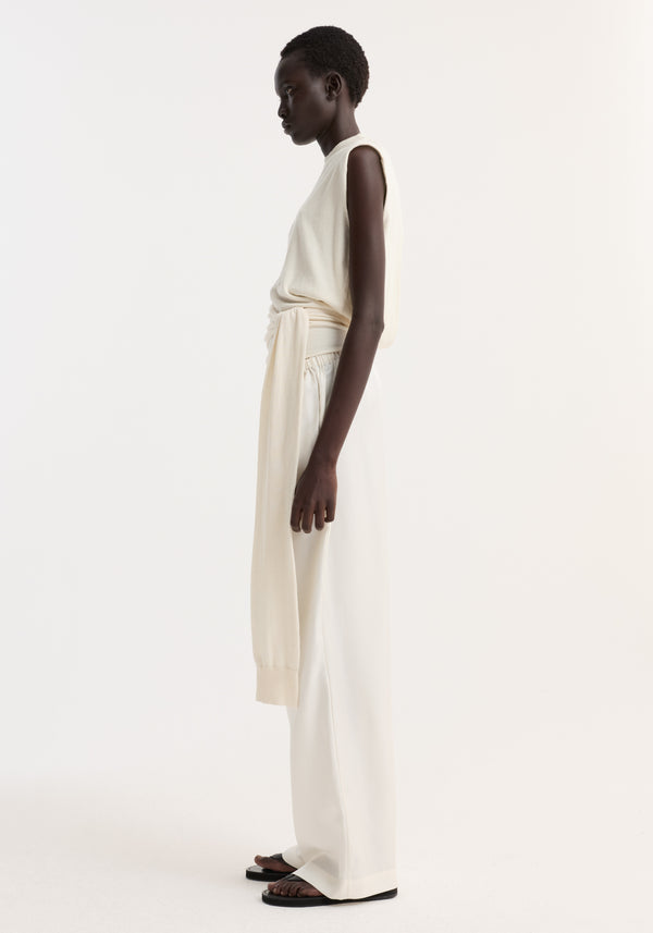A person wears an Open back knitted wrap top | off white, featuring a sleeveless design, open back, wrap waist, and sash-style sleeve extensions.