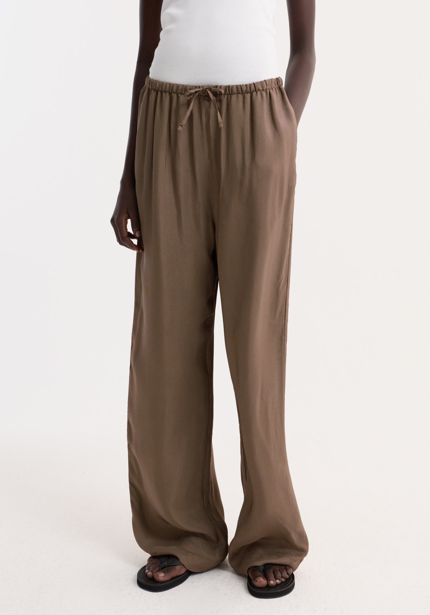 Person wearing Relaxed fit trousers | brown mini check, featuring a loose, wide-leg design with an elasticated drawstring waist and subtle check pattern, paired with black flip-flops.
