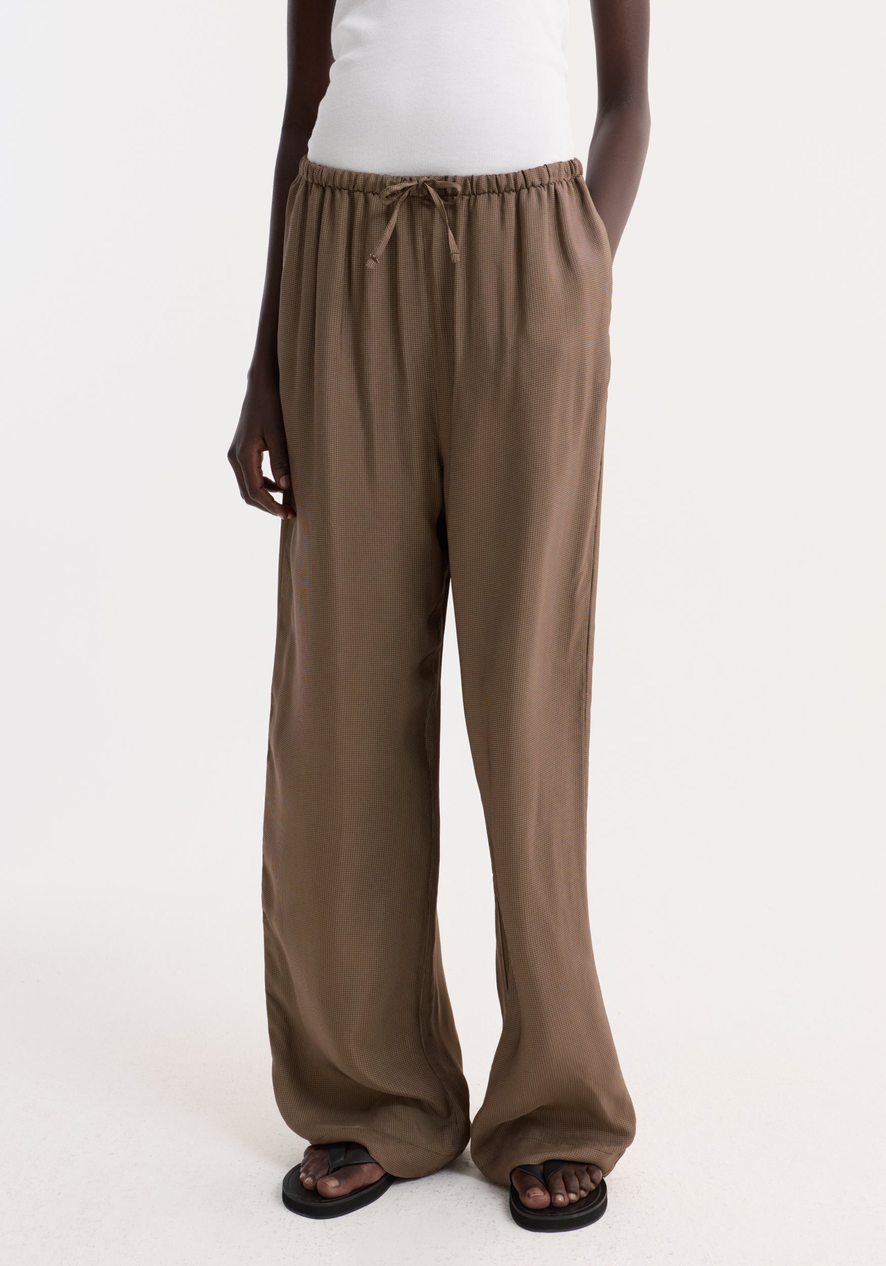 Person wearing Relaxed fit trousers | brown mini check, featuring a loose, wide-leg design with an elasticated drawstring waist and subtle check pattern, paired with black flip-flops.