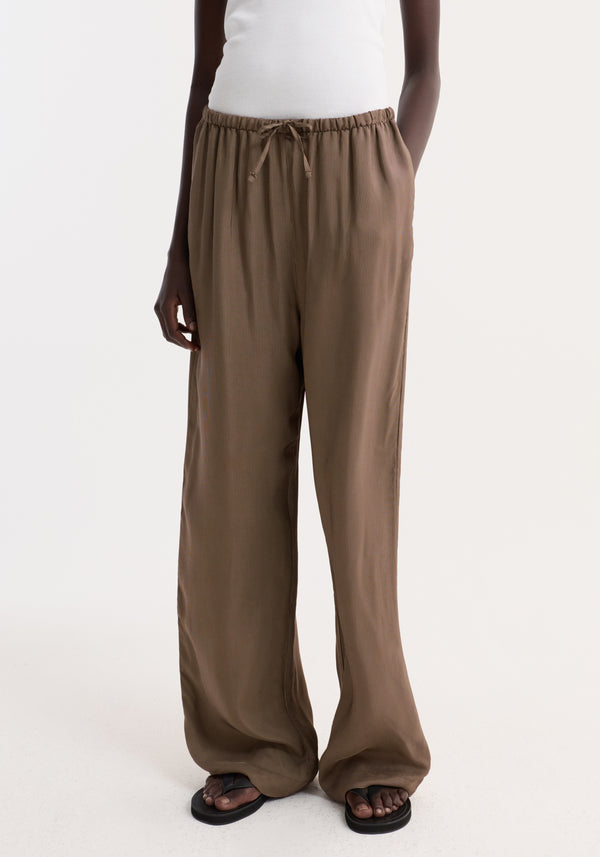 Person wearing Relaxed fit trousers | brown mini check, featuring a loose, wide-leg design with an elasticated drawstring waist and subtle check pattern, paired with black flip-flops.