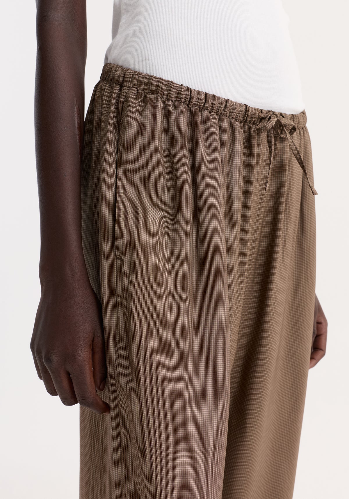 Relaxed fit trousers in brown mini check, featuring a gathered elasticized drawstring waistband. The image highlights the trousers' loose fit and ribbed top detail.