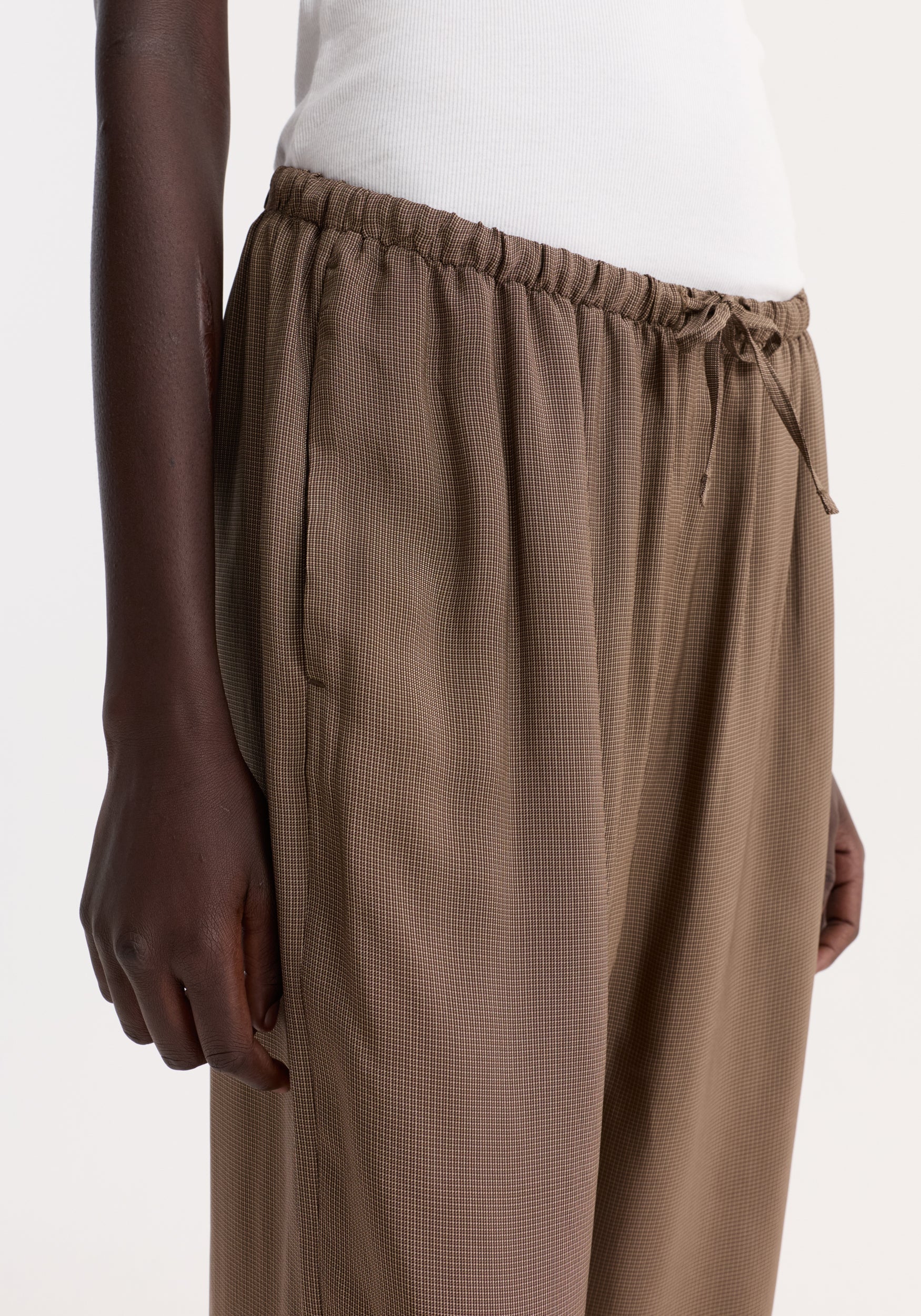 Relaxed fit trousers in brown mini check, featuring a gathered elasticized drawstring waistband. The image highlights the trousers' loose fit and ribbed top detail.