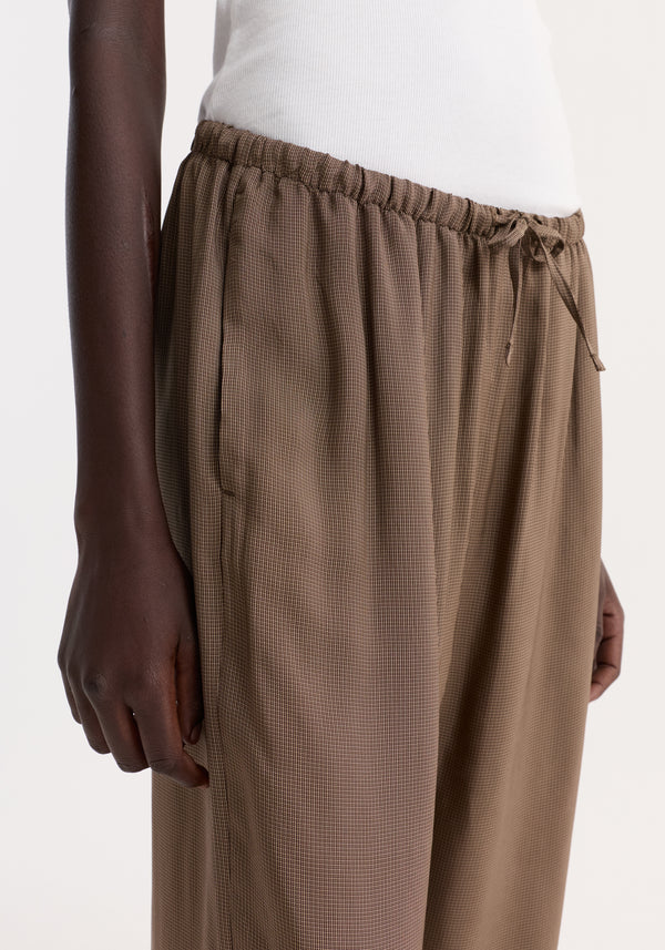 Relaxed fit trousers in brown mini check, featuring a gathered elasticized drawstring waistband. The image highlights the trousers' loose fit and ribbed top detail.