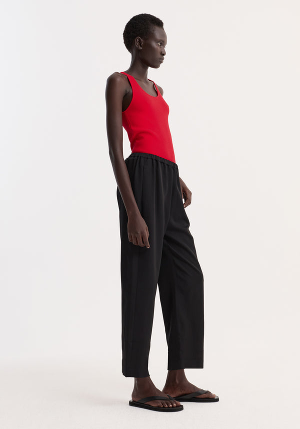 Model wearing cropped wide leg trousers in black, featuring an elasticated waist, ankle-length fit, and side pockets, paired with a bright red sleeveless top.