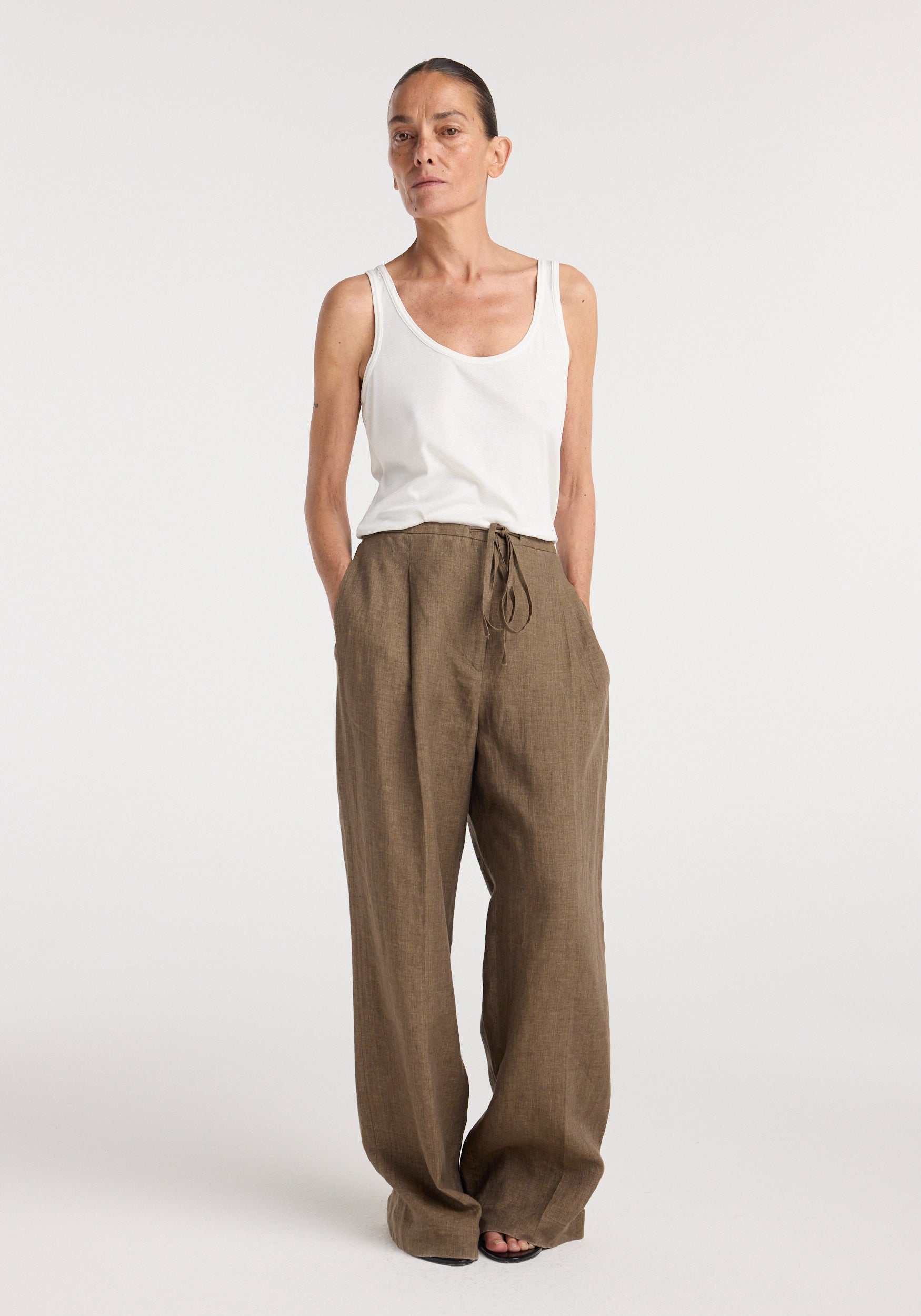 Woman wearing relaxed drawstring trousers | brown melange paired with a white sleeveless top, standing against a plain white background with hands in pockets.