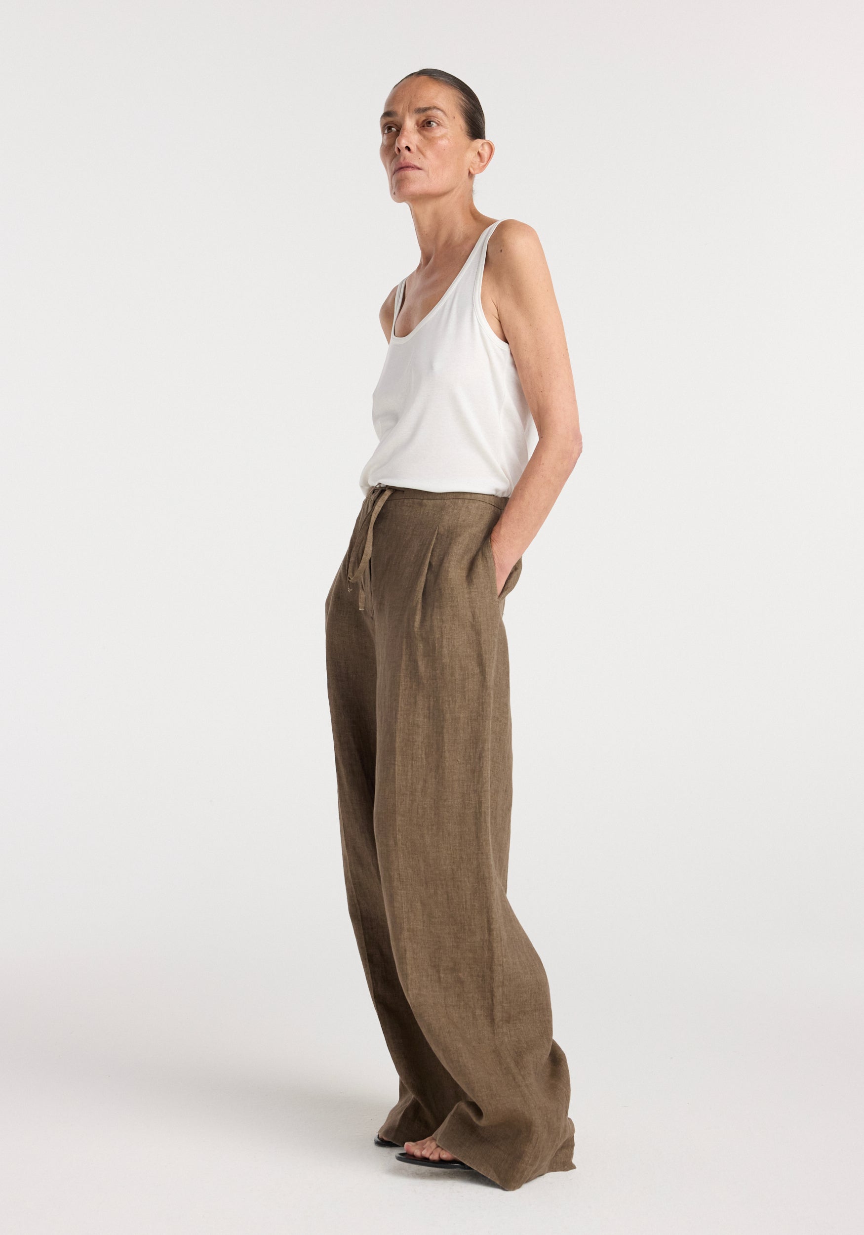 Woman wearing relaxed drawstring trousers | brown melange paired with a white sleeveless top, standing with hands in pockets against a plain background.