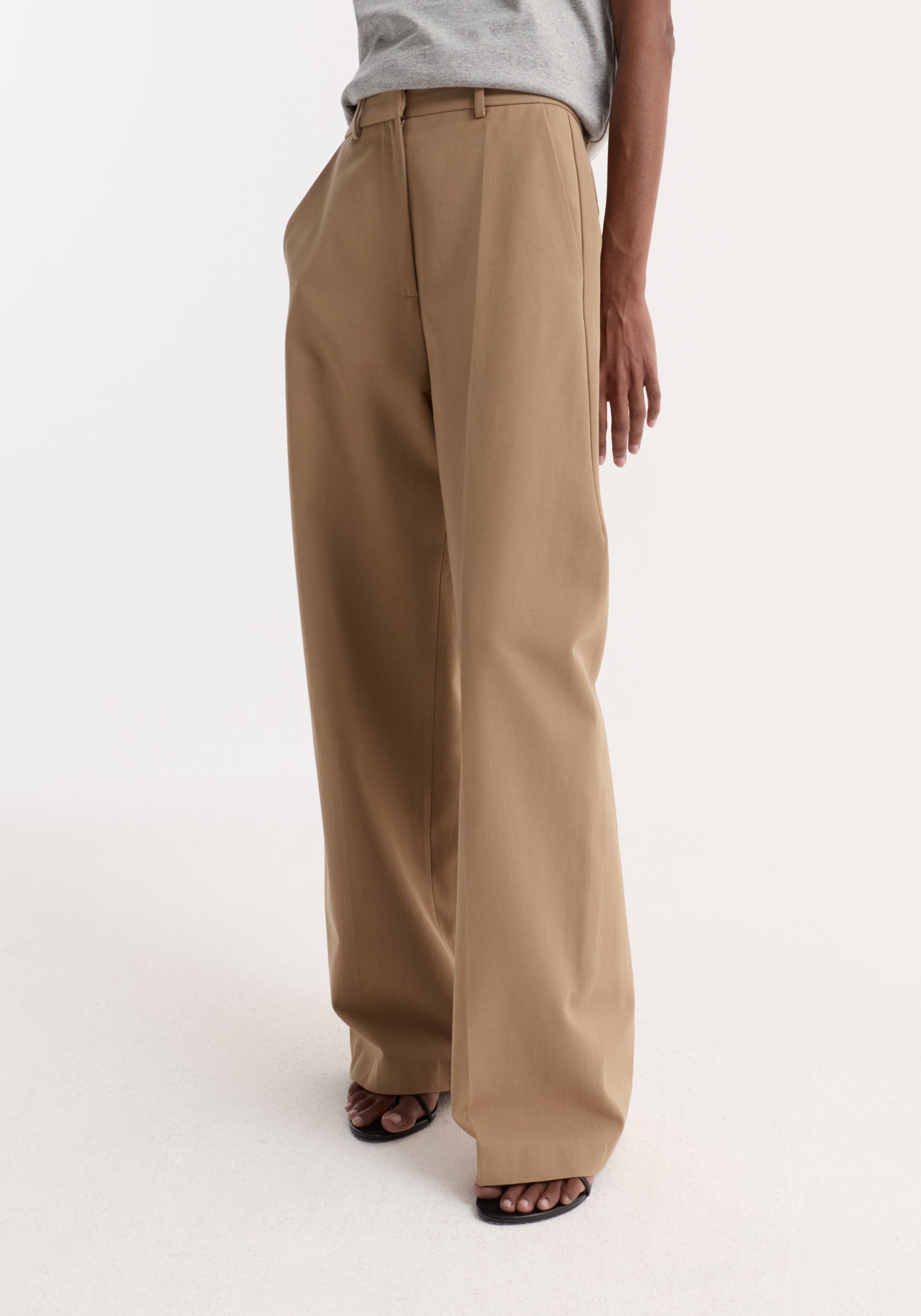 Model wearing Classic loose fit chino | beige, showcasing high-waisted, wide-leg trousers with side pockets; paired with a tucked-in gray T-shirt and black thong sandals.