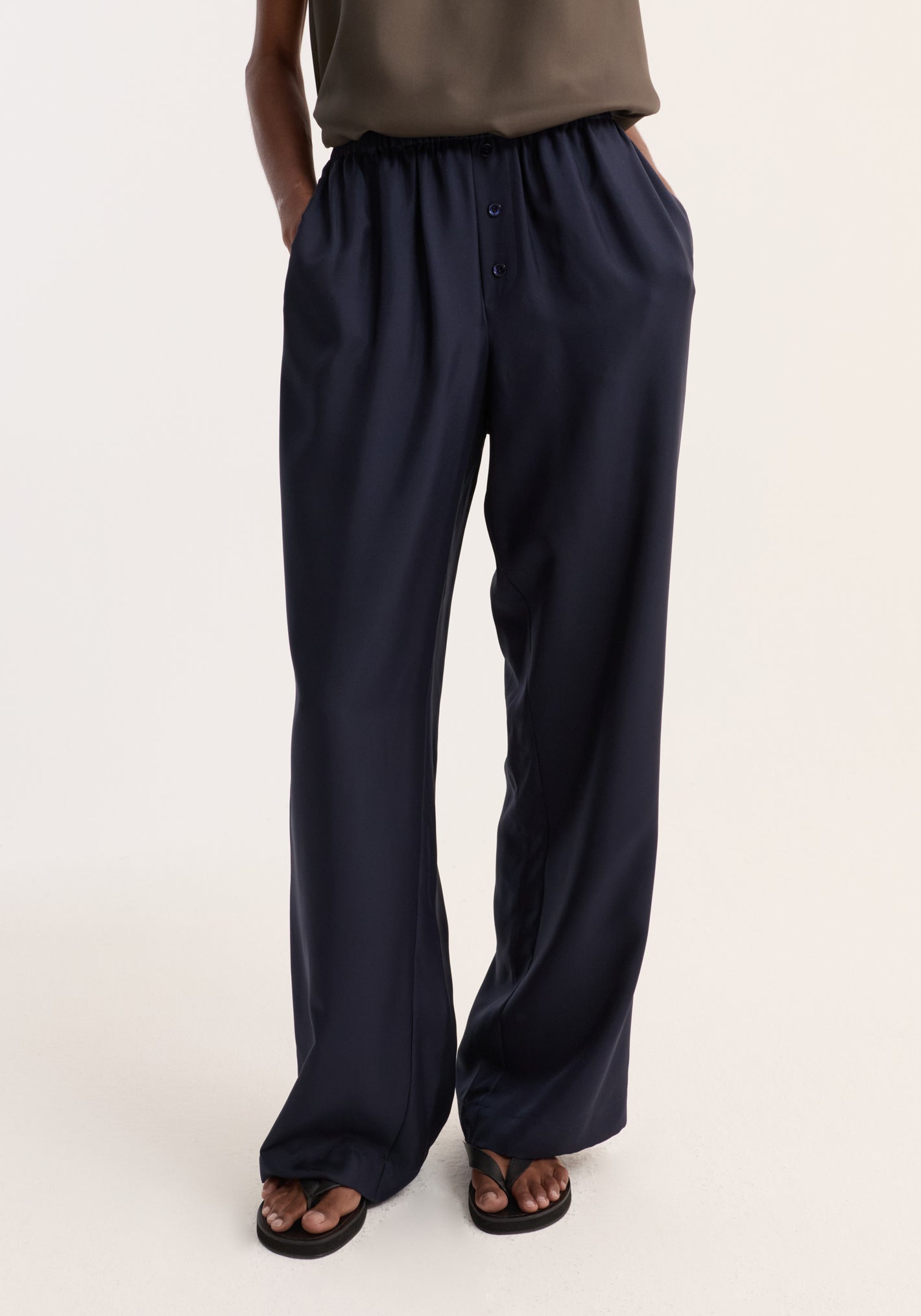 Relaxed silk trousers | night, featuring a wide leg and elastic waistband with two-button detail, modeled with hands in pockets and black flip-flops.