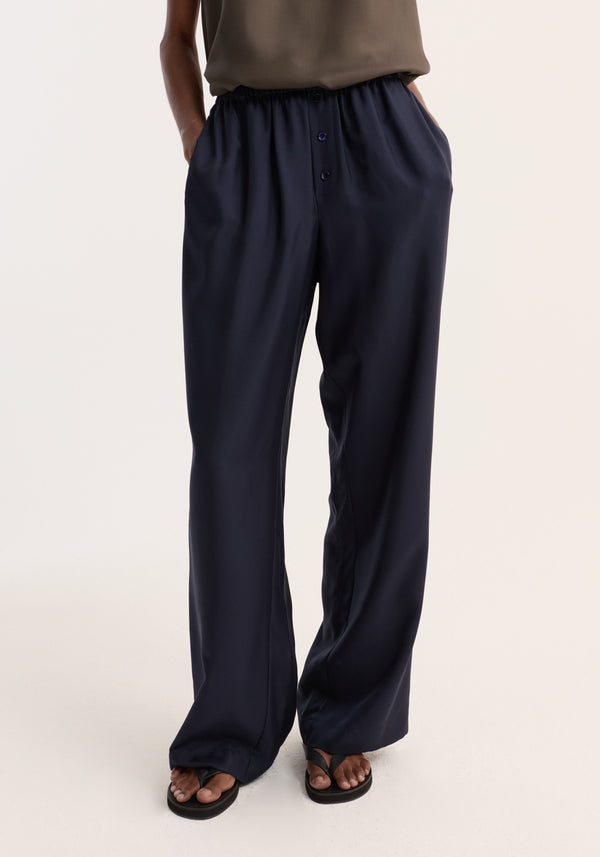 Relaxed silk trousers | night, featuring a wide leg and elastic waistband with two-button detail, modeled with hands in pockets and black flip-flops.