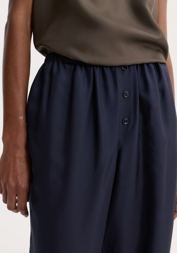 The image shows Relaxed silk trousers | night, featuring a high-waisted, wide-leg design with an elasticated waistband and faux fly with button detailing.