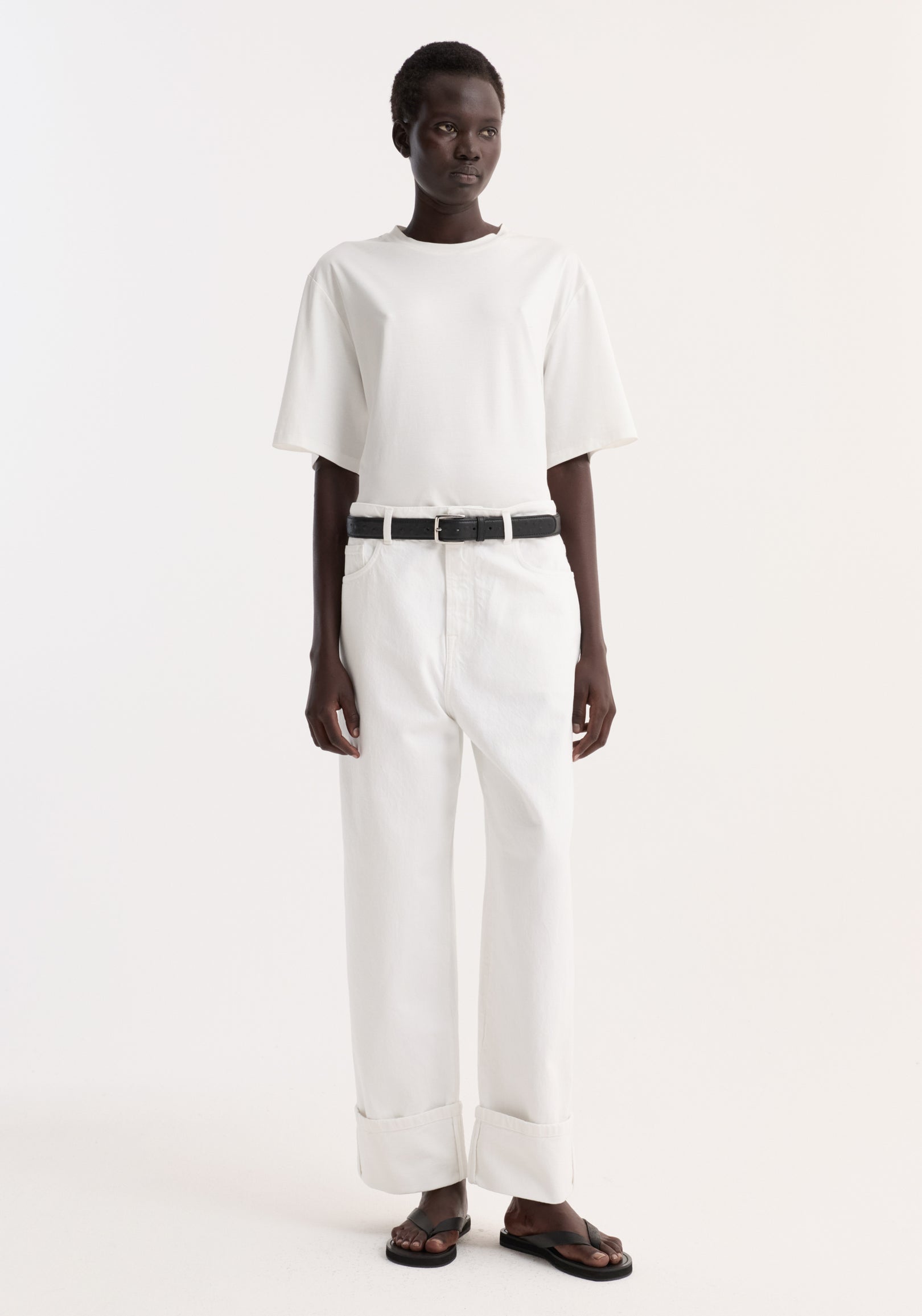 Model wearing Baggy fit turn-up denim in optic white, featuring wide-leg, low-rise fit with rolled cuffs, paired with a black belt and sandals.