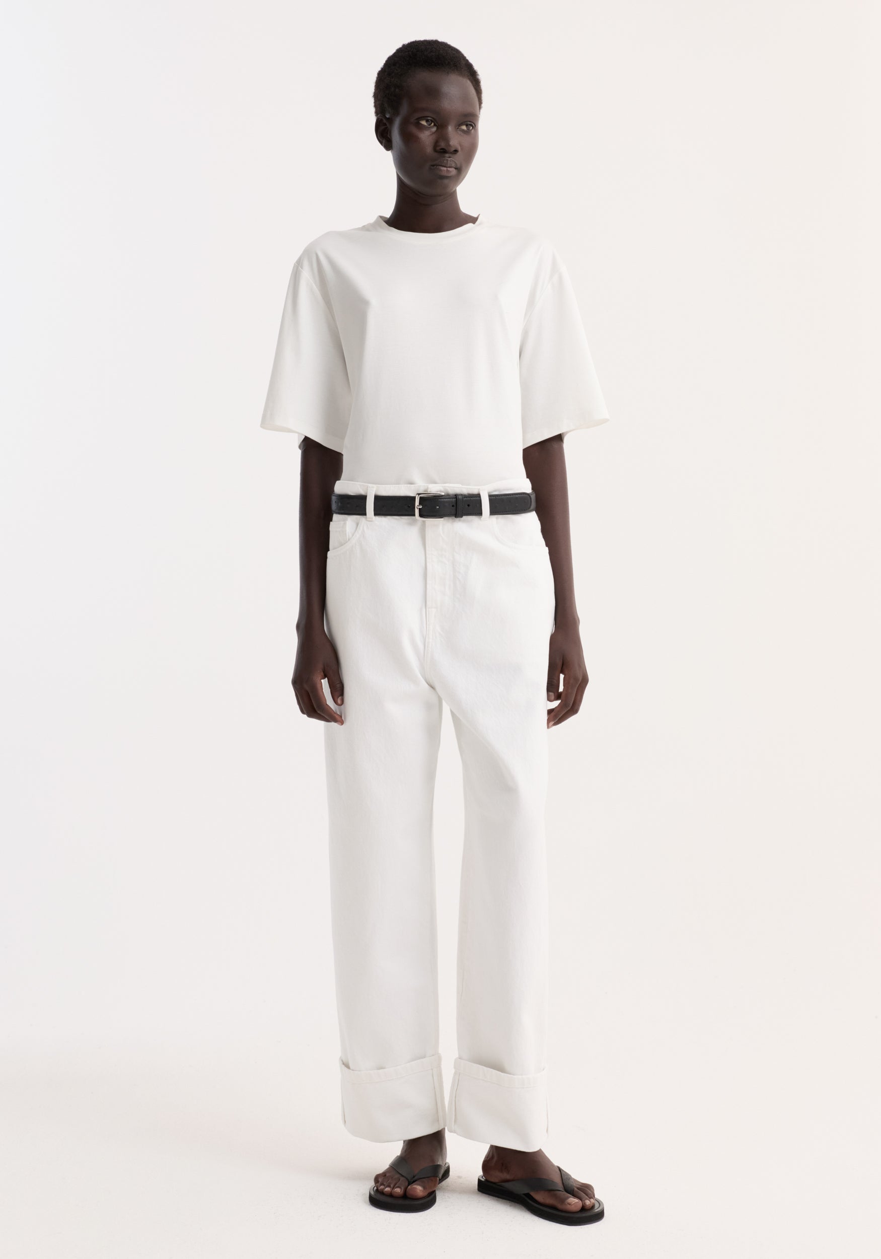 Model wearing Baggy fit turn-up denim in optic white, featuring wide-leg, low-rise fit with rolled cuffs, paired with a black belt and sandals.