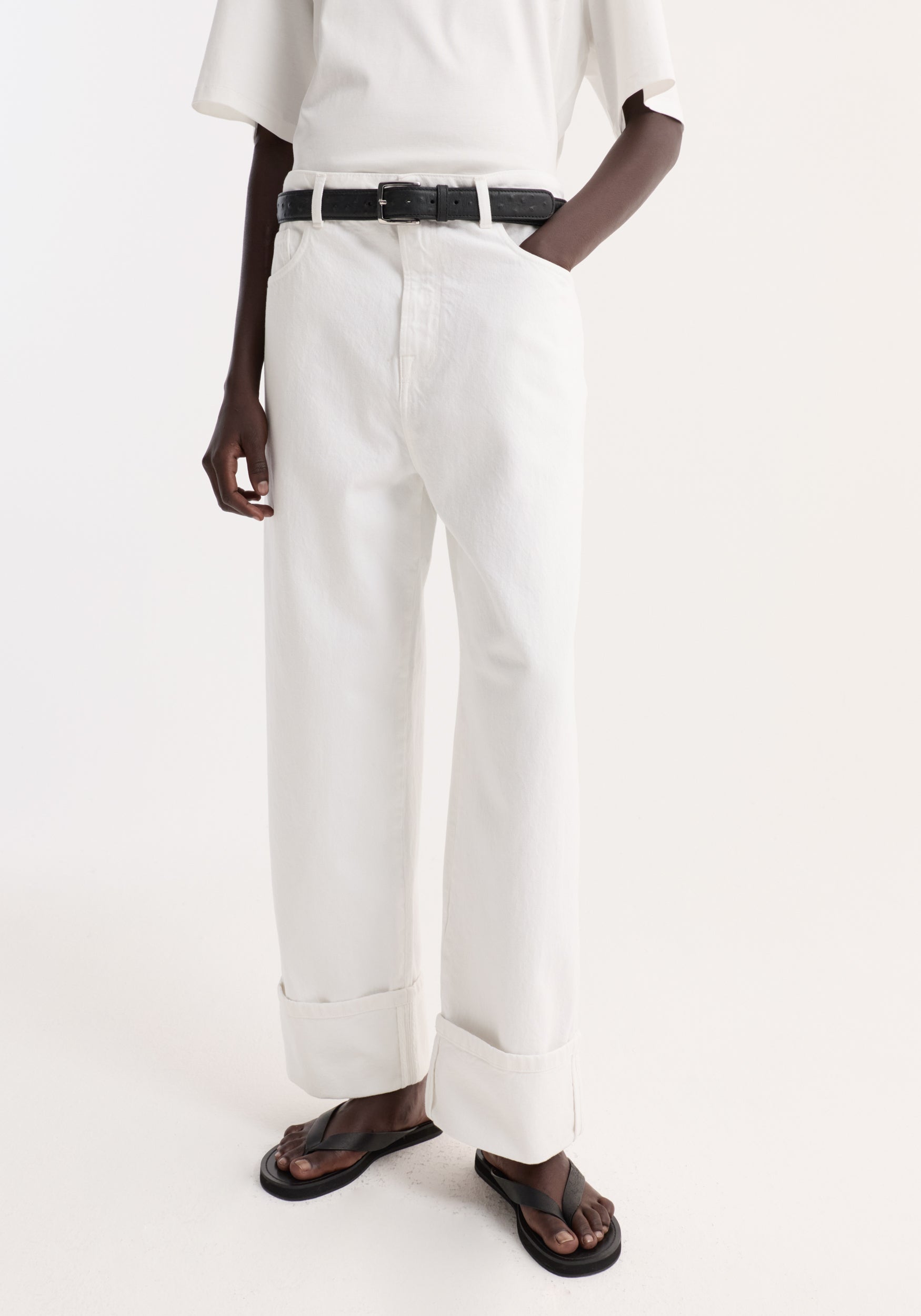 Baggy fit turn-up denim in optic white; high-waisted, wide-leg trousers with rolled cuffs, black leather belt, and black sandals. T-shirt tucked in.
