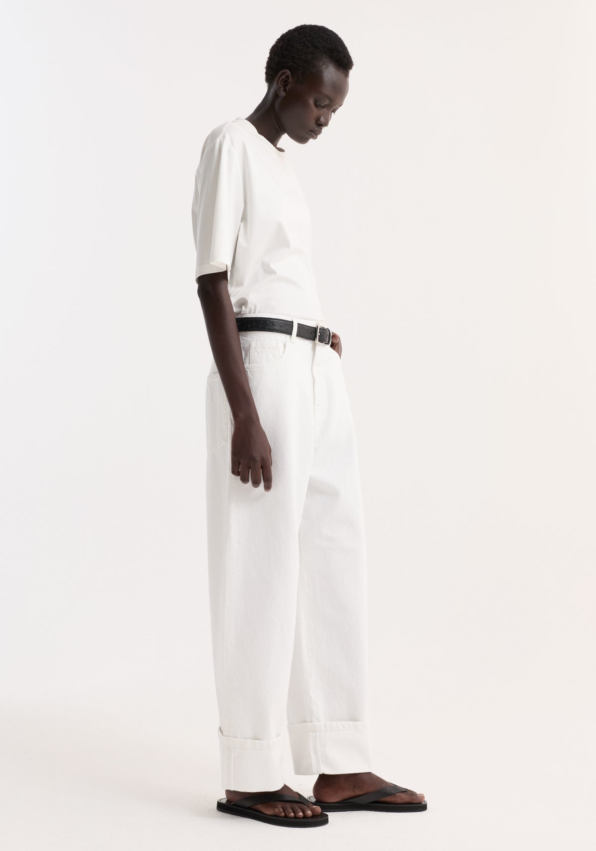 Model in profile wears Baggy fit turn-up denim in optic white, featuring wide-leg, low-waist design with prominent cuff, paired with black belt and sandals.