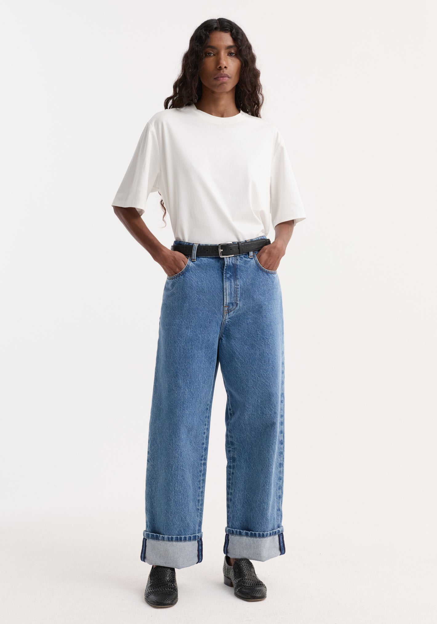 Person wearing Turn-up baggy denim | heritage blue, featuring high-waisted, wide-leg, turned-up hems, with a black belt and black woven slip-on shoes.
