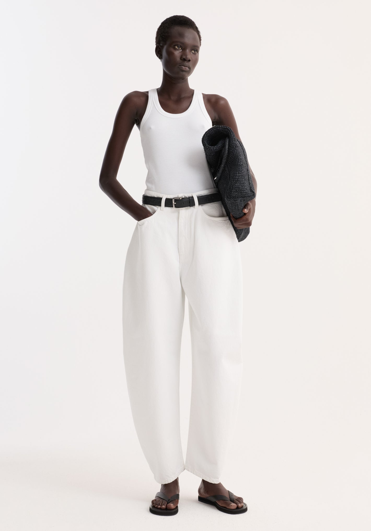 Model wearing Sculptural denim trousers in optic white, featuring high waist, balloon leg, relaxed fit, paired with a white tank top and black sandals.
