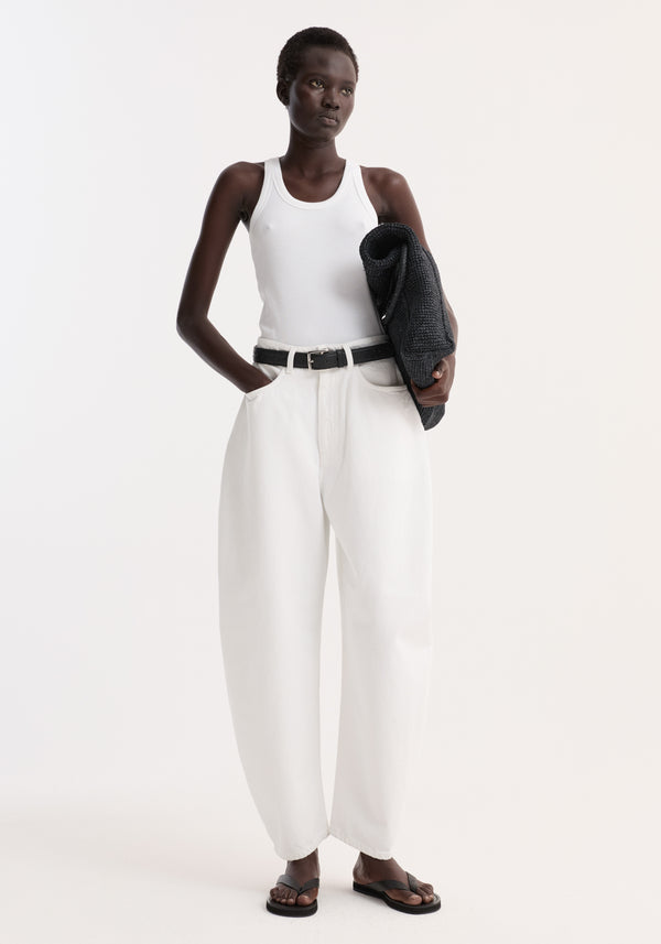 Model wearing Sculptural denim trousers in optic white, featuring high waist, balloon leg, relaxed fit, paired with a white tank top and black sandals.