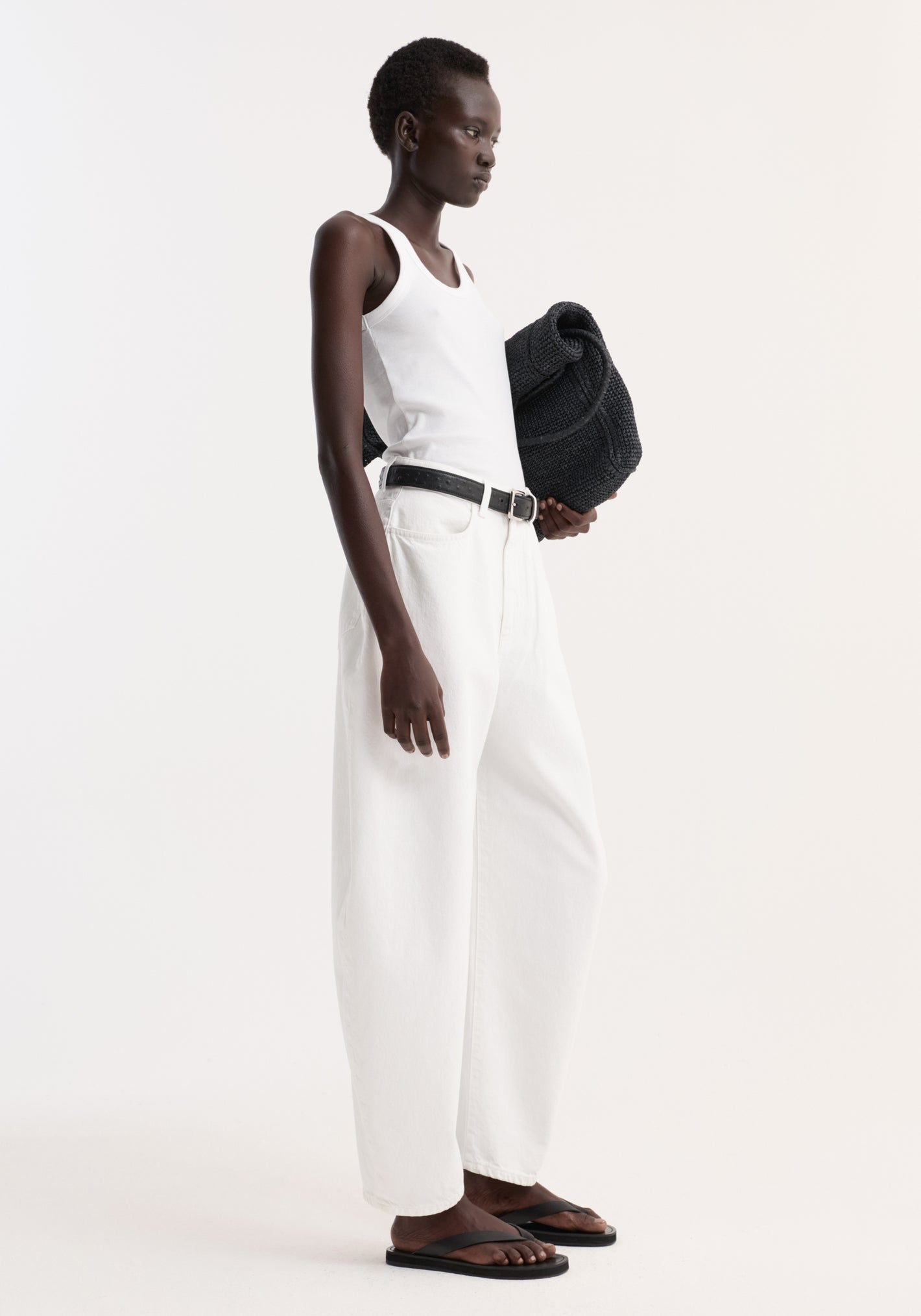 Model wearing Sculptural denim trousers | optic white, showcasing high-waisted, balloon leg design, paired with a white tank, black belt, and sandals, holding a black item.