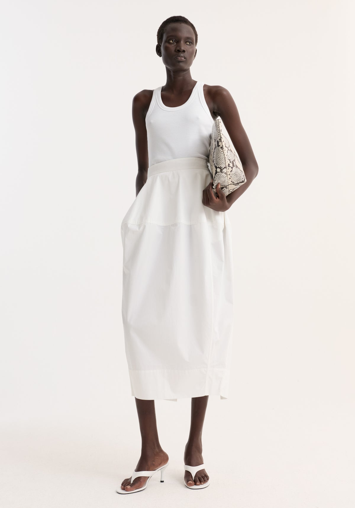Model wearing sculptural poplin skirt in optic white, featuring a high waist, seam detailing, and back slit, paired with white tank top and sandals.