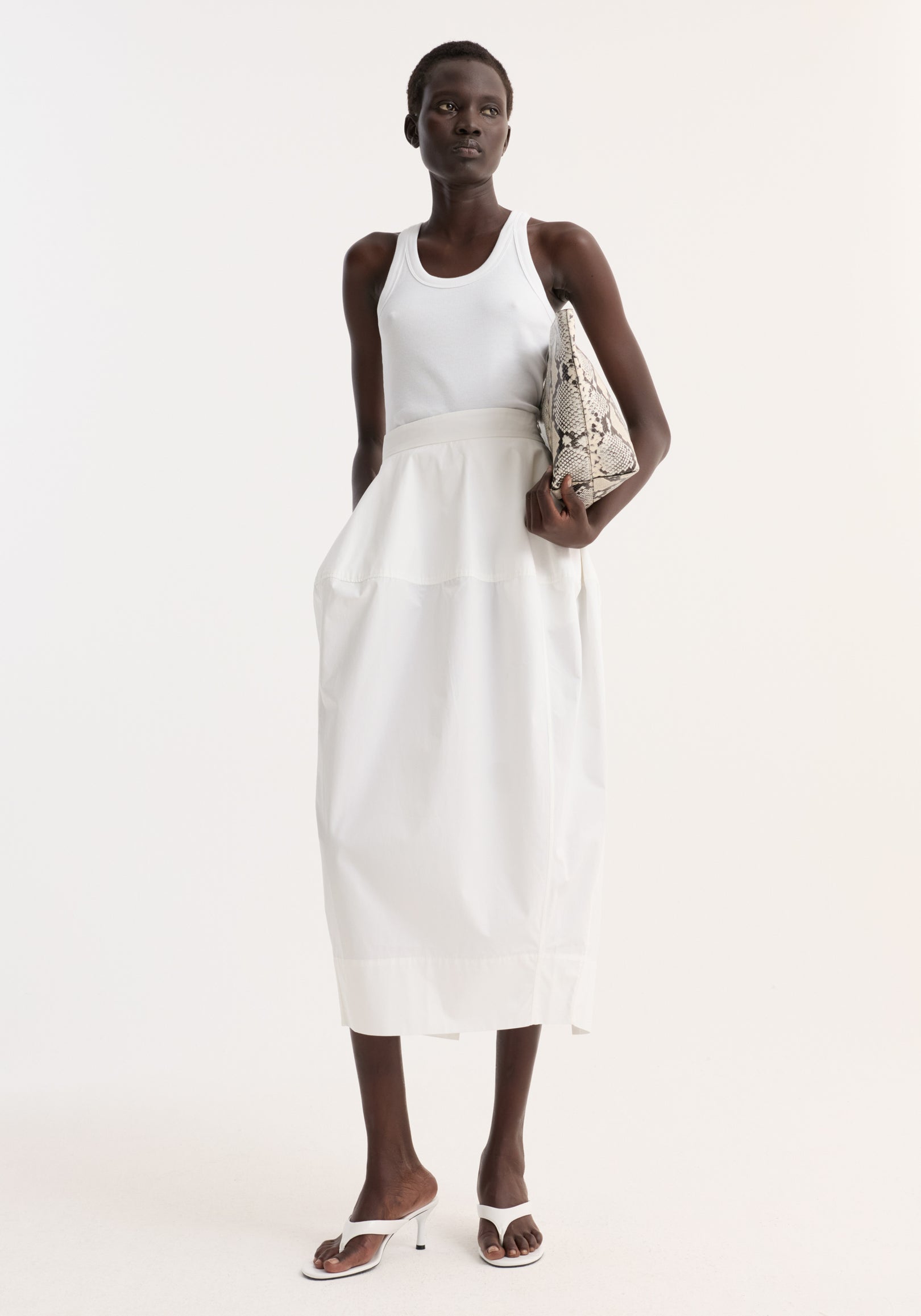 Model wearing sculptural poplin skirt in optic white, featuring a high waist, seam detailing, and back slit, paired with white tank top and sandals.