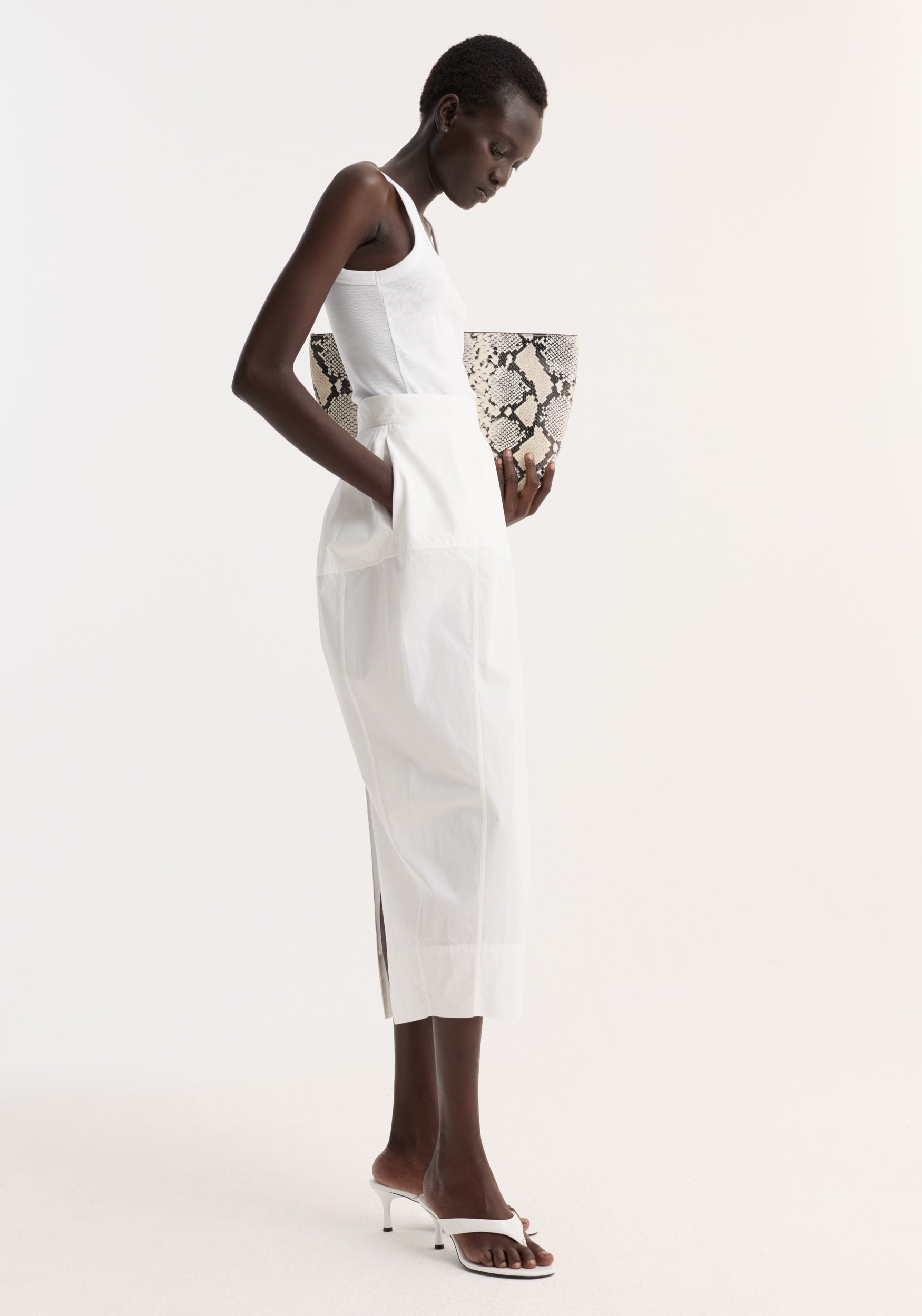 Model in Sculptural poplin skirt | optic white, showcasing high waist and seam detailing, paired with a snakeskin tote and white mules against a plain background.