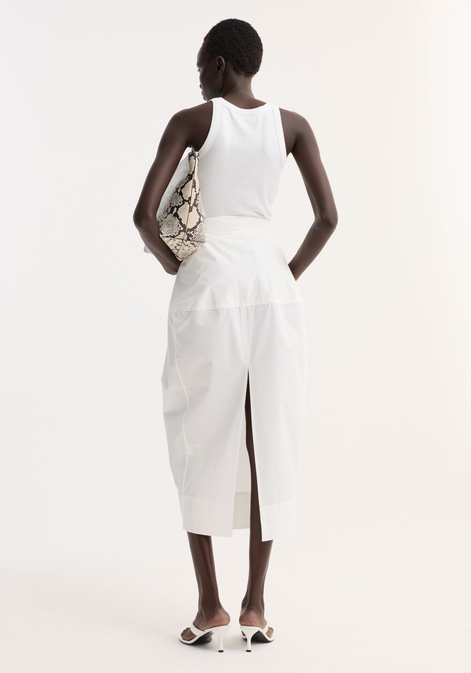 Woman in a white ribbed tank and sculptural poplin skirt, optic white, with seam detailing and back slit, holding a snakeskin-print pouch under her arm.