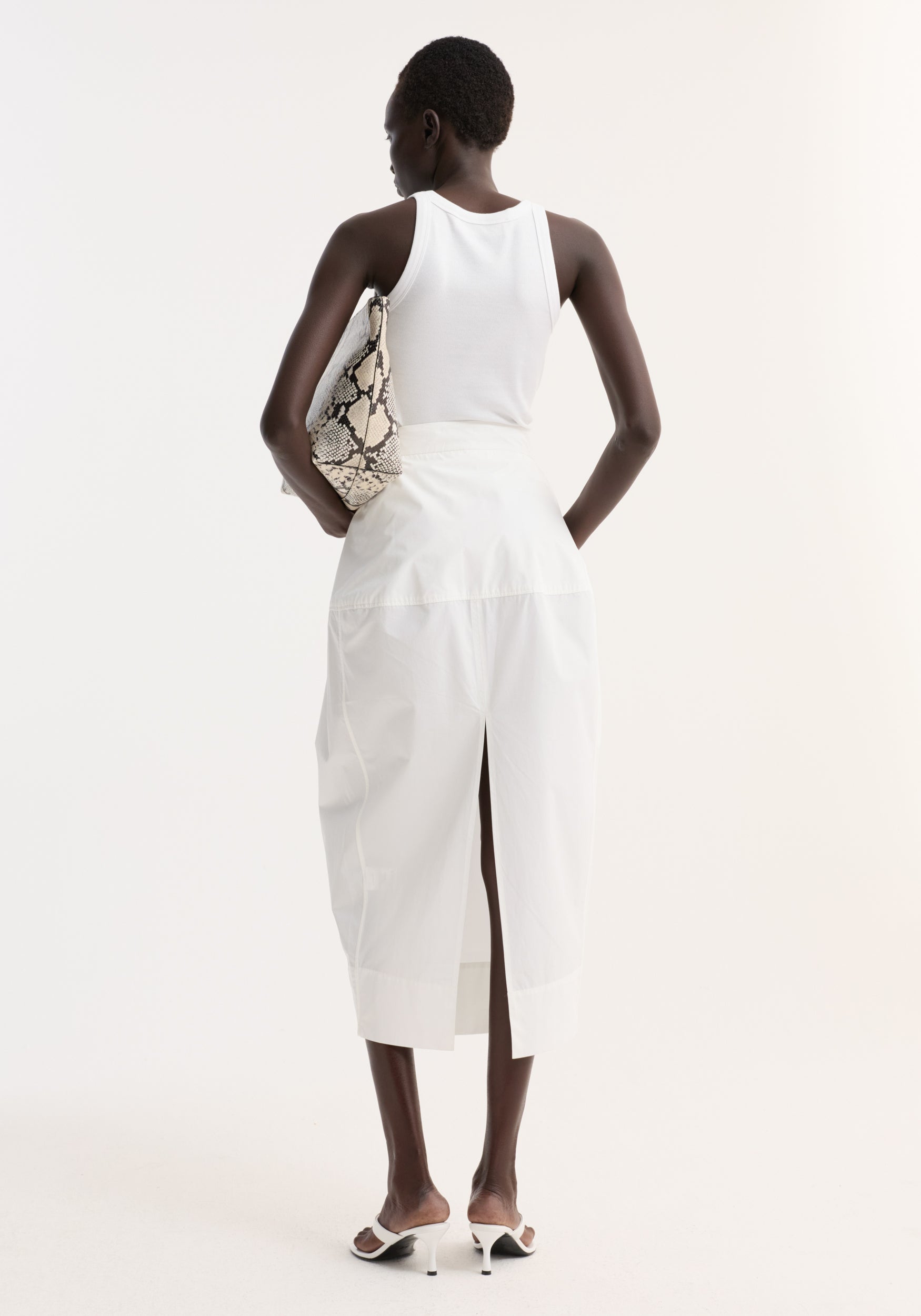 Woman in a white ribbed tank and sculptural poplin skirt, optic white, with seam detailing and back slit, holding a snakeskin-print pouch under her arm.