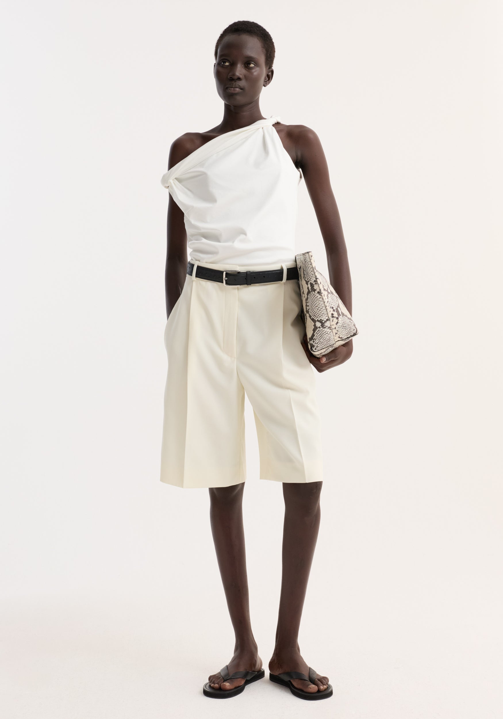 Model wearing Tailored single pleat shorts | off white with a relaxed fit, side pockets, and a black leather belt, paired with a one-shoulder top.